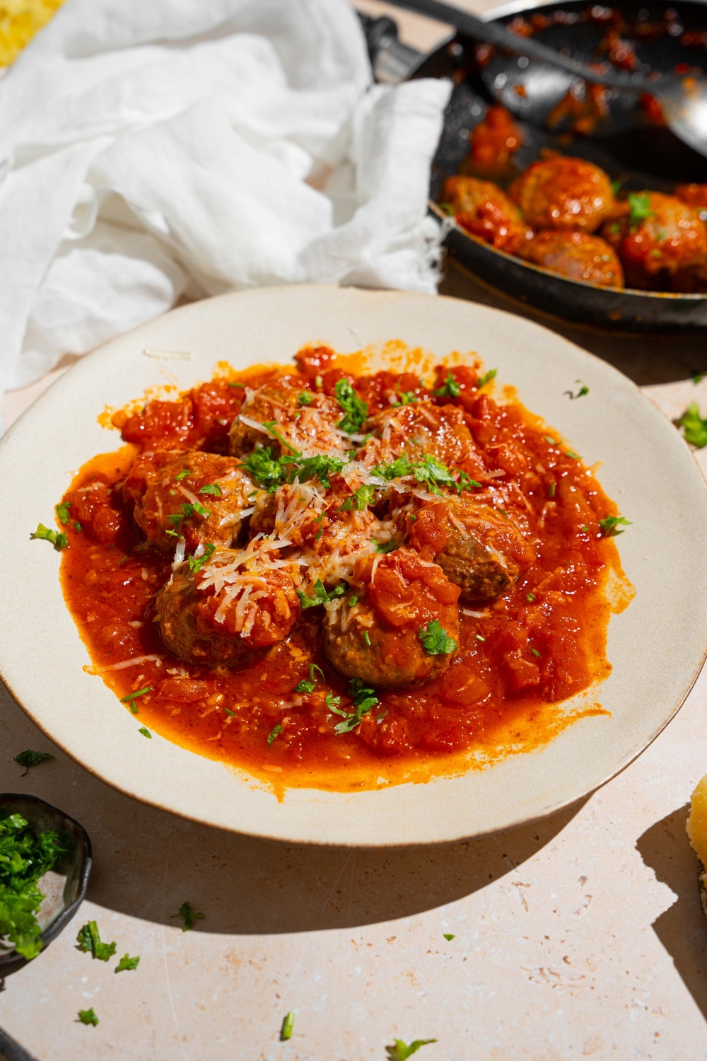 A white plate with frozen meatballs cooked in tomato sauce topped with grated cheese and fresh parsley. The plate is on a tan counter with a white cloth napkin and skillet of meatballs.