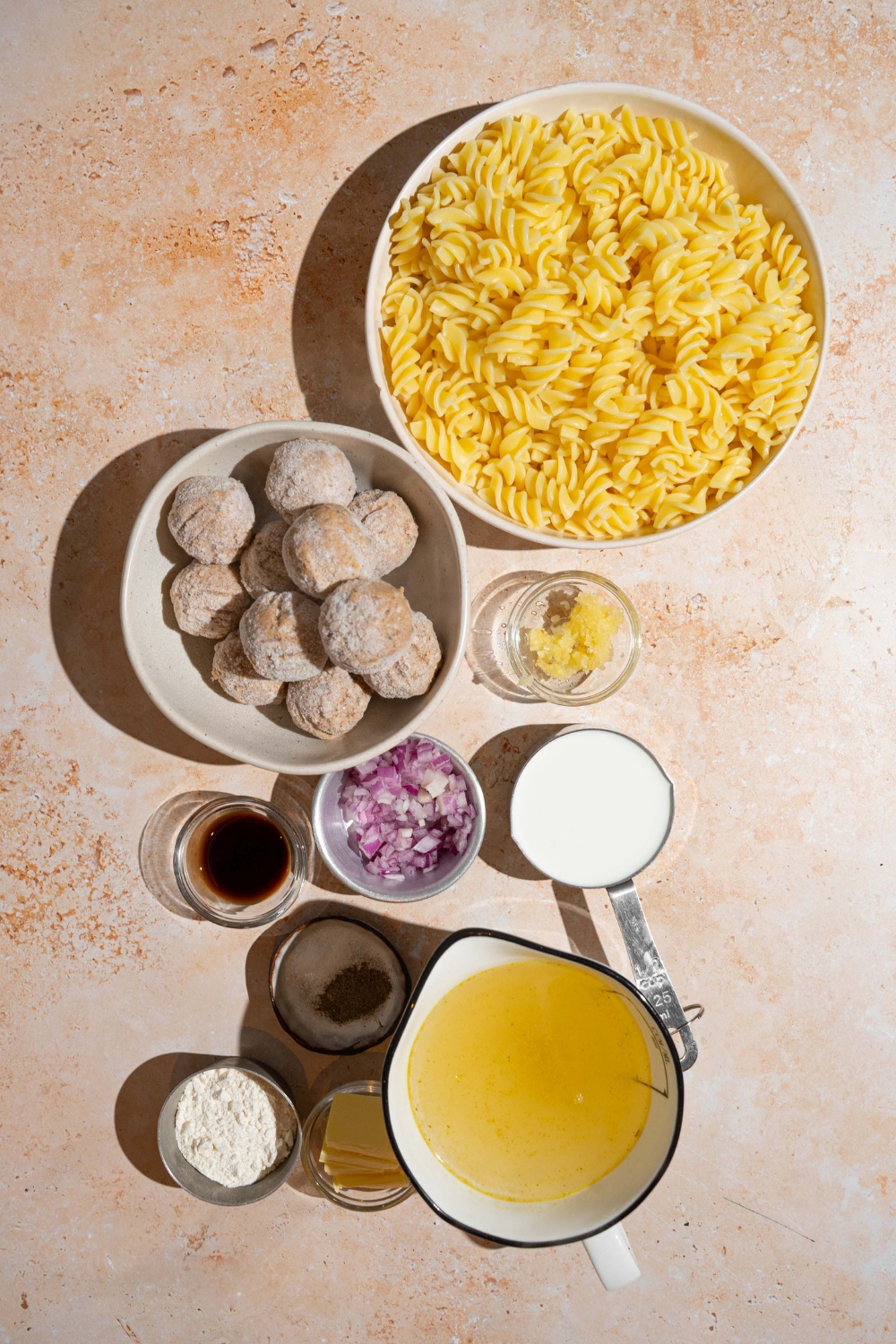 An overhead shot of several bowls in various sizes containing ingredients to make Swedish meatballs including frozen meatballs, rotini pasta, cream, chicken stock, Worcestershire sauce, butter, and seasonings.