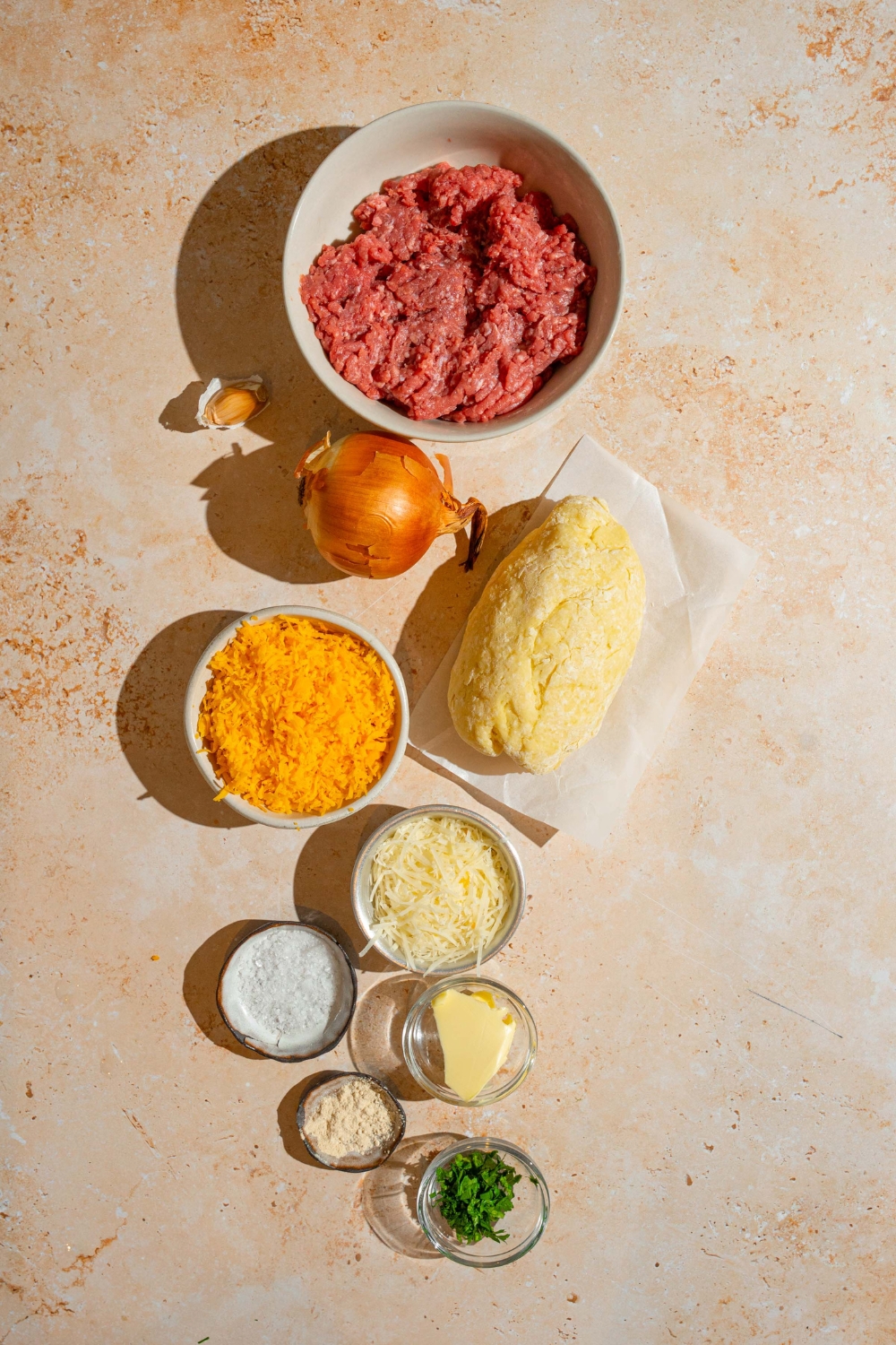 An overhead shot of several bowls in various sizes containing ingredients to make garlic parmesan cheeseburger bites including ground beef, shredded cheddar cheese, biscuit dough, onion, butter, shredded cheese, and more.