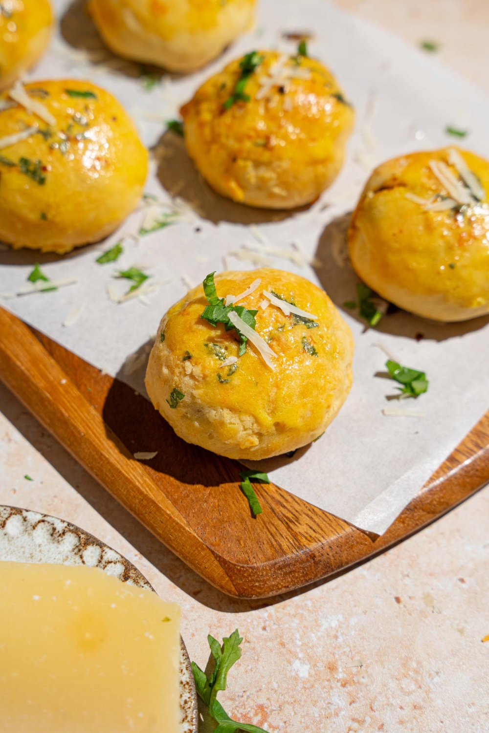 A wooden board lined with parchment paper with several garlic parmesan cheeseburger bombs garnished with fresh parsley and grated cheese. The board is on a tan counter with a wedge of cheese.