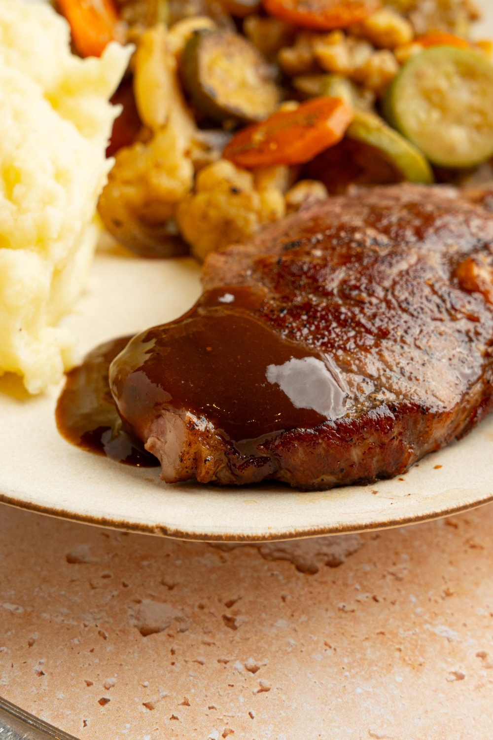 A white plate with mashed potatoes, roasted vegetables, and steak topped with homemade A1 steak sauce. The plate is on a tan counter with a condiment jar of steak sauce and skillet.