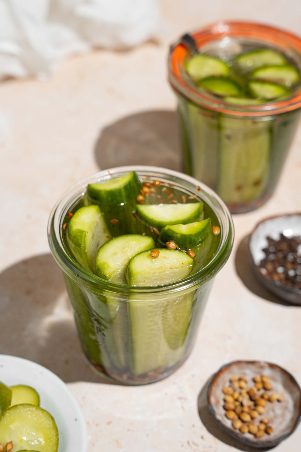 An open jar of homemade Claussen pickles on a tan counter with an additional sealed jar of pickles, plates of seasonings, and small bowl of sliced pickles.