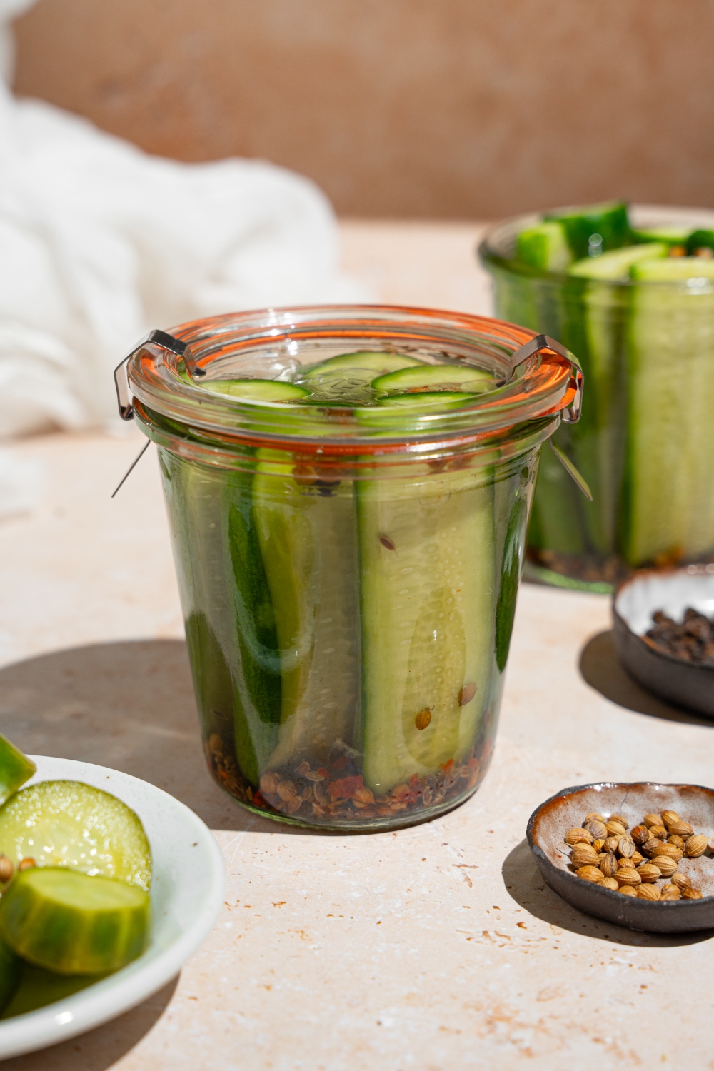 A sealed jar of homemade Claussen pickles on a tan counter with an additional open jar of pickles, plates of seasonings, and small bowl of sliced pickles. There is a fork taking a pickle spear from the jar.