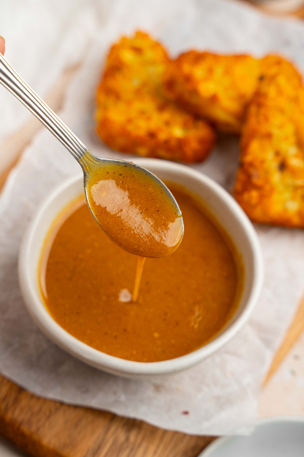 A spoon drizzling McDonald's golden sauce into a white bowl of sauce. The bowl is on a wooden board lined with parchment paper with crispy hashbrowns.