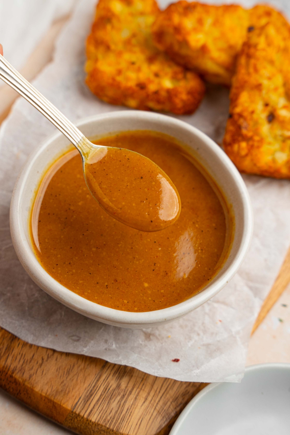 A spoon with McDonald's golden sauce over a white bowl of sauce. The bowl is on a wooden board lined with parchment paper with crispy hashbrowns.