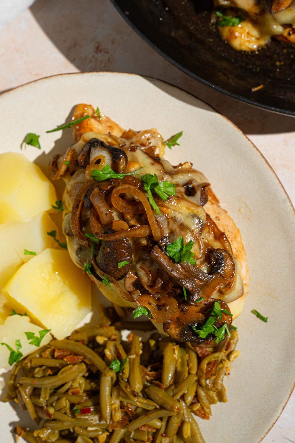 A piece of Texas Roadhouse smothered chicken on a white plate with cubed potatoes and green beans. The plate is on garnished with fresh parsley. The plate is on a tan counter with a skillet with additional chicken.