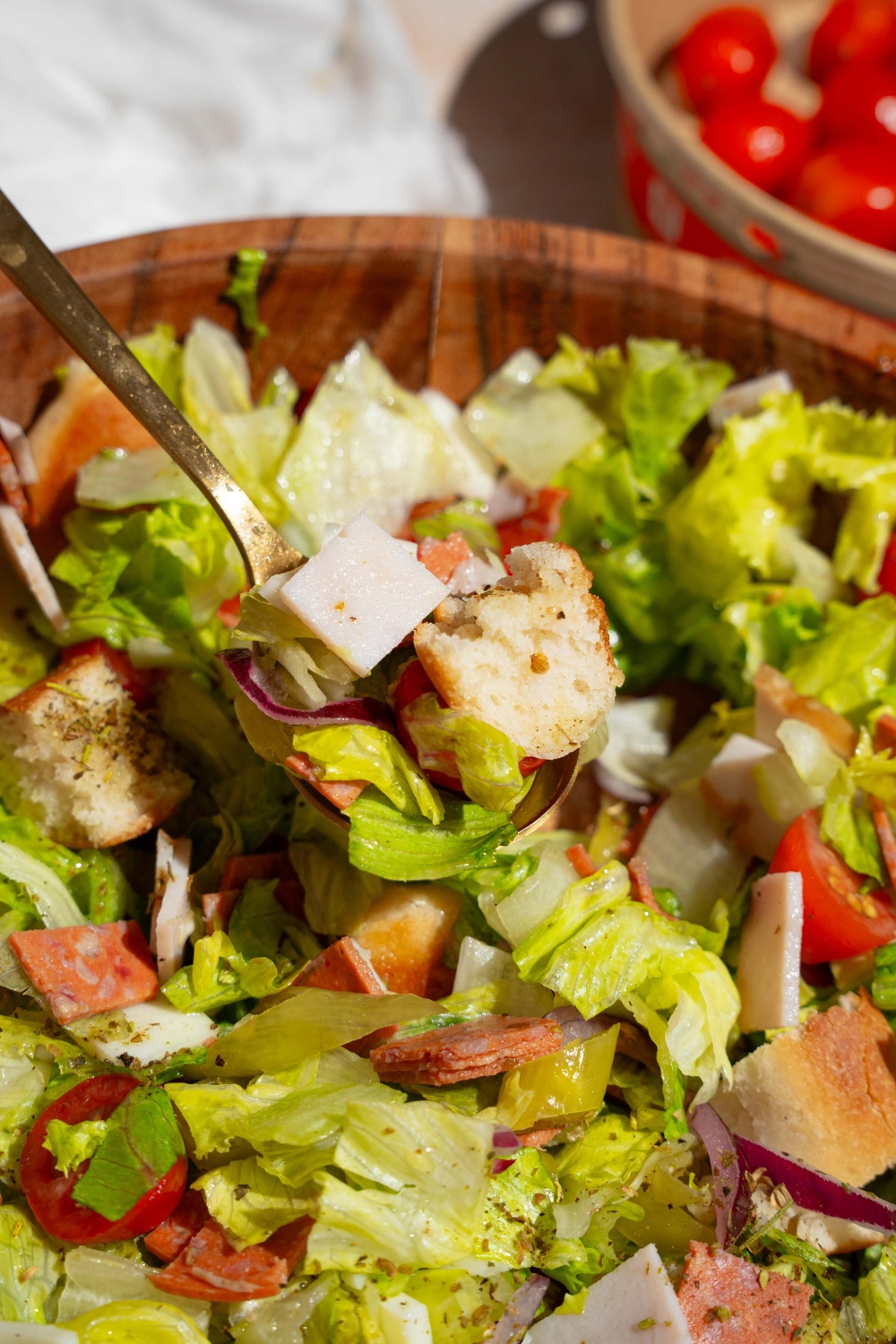 A wooden salad bowl of copycat Jersey Mike's sub in a tub including mixed lettuce, tomatoes, provolone cheese, diced meats, and bread croutons. There is a fork taking a bite from the salad.