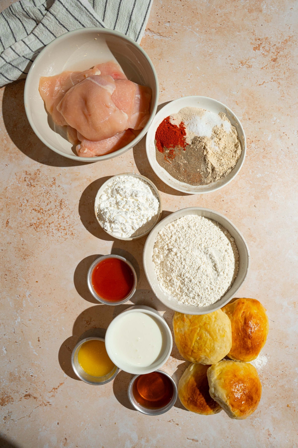 An overhead shot of several bowls in various sizes containing ingredients to make hot honey chicken sandwiches including chicken breasts, buns, buttermilk, hot sauce, honey, butter, cornstarch, flour, and seasonings.