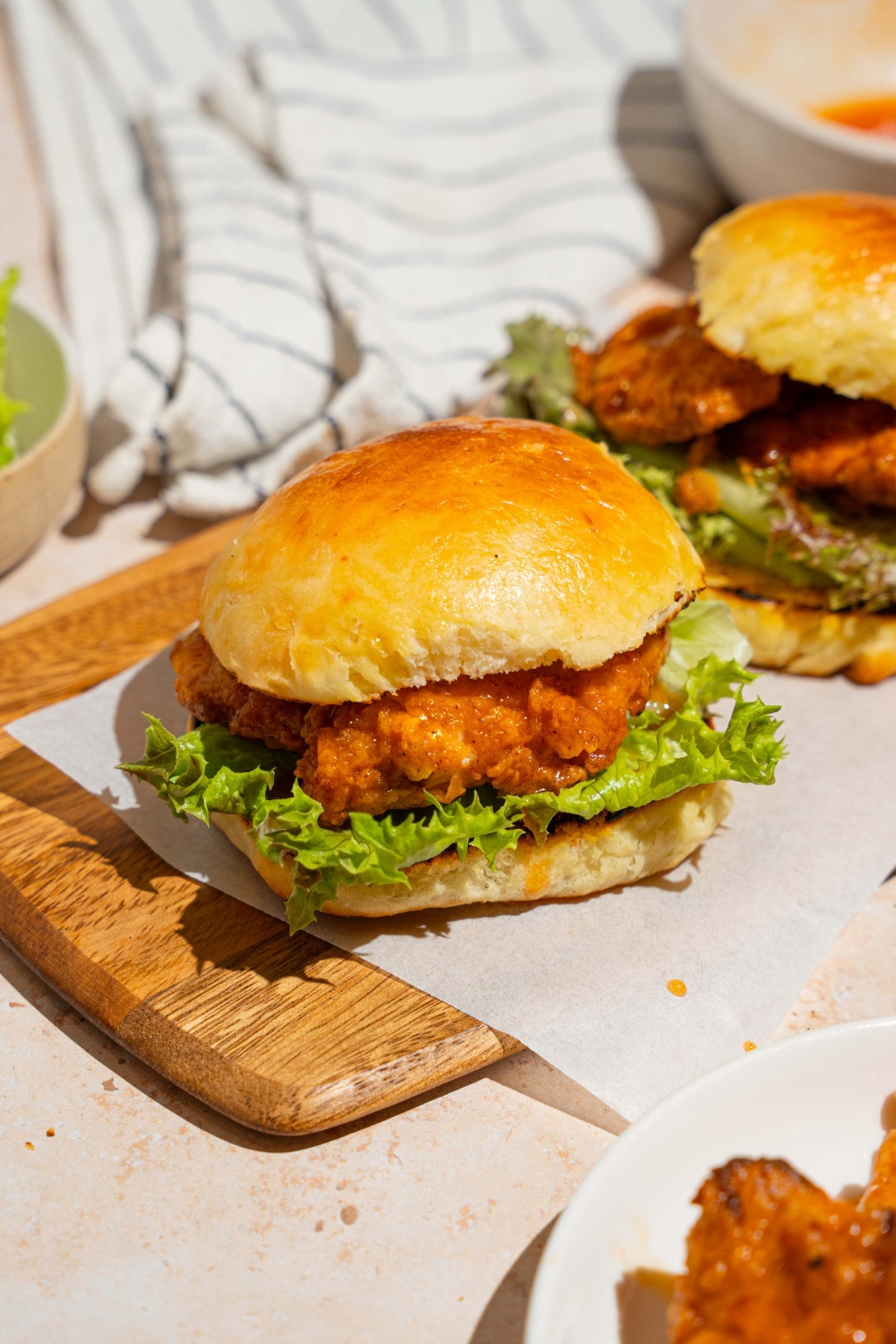 A wooden board lined with parchment paper with two hot honey chicken sandwiches. The board is on a tan counter with a white striped napkin.