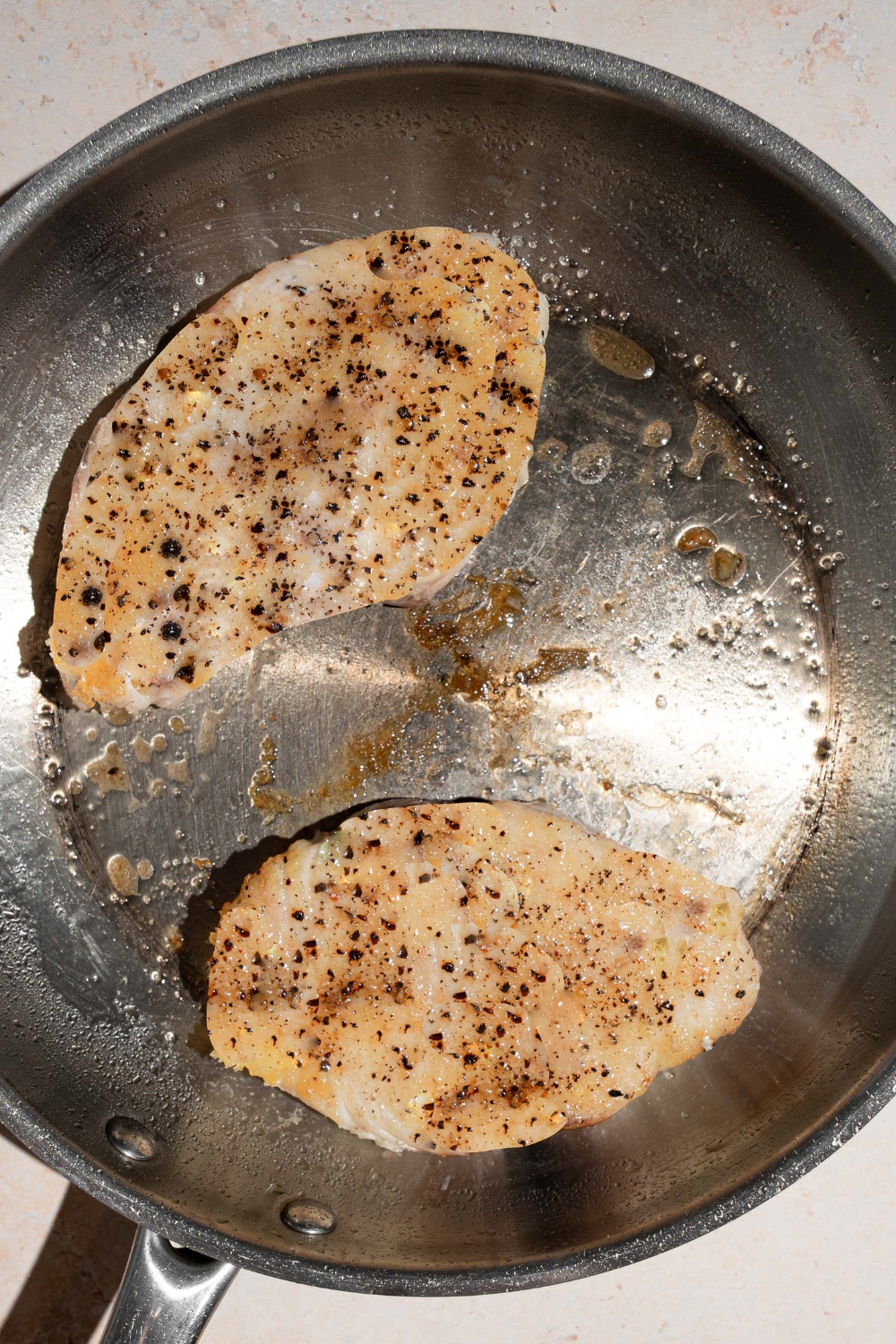 A skillet with two pan seared swordfish fillets cooking in oil. The skillet is on a tan counter.