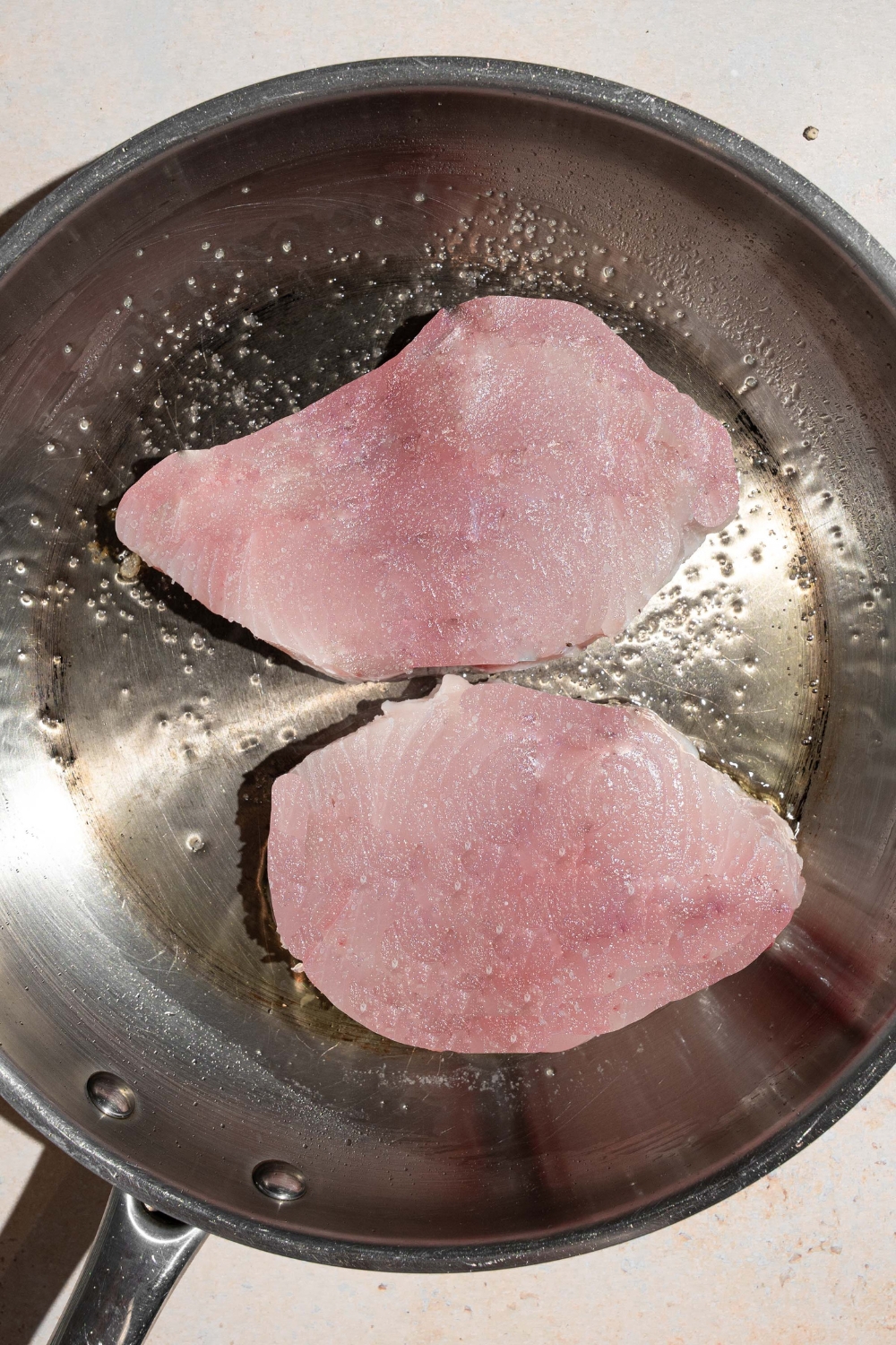A skillet with two uncooked swordfish fillets cooking in oil and seasonings.