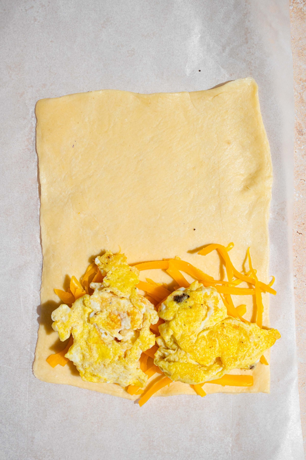 An open puff pastry sheet with scrambled egg and shredded cheddar cheese on the bottom half. The pastry is on a sheet of parchment paper.