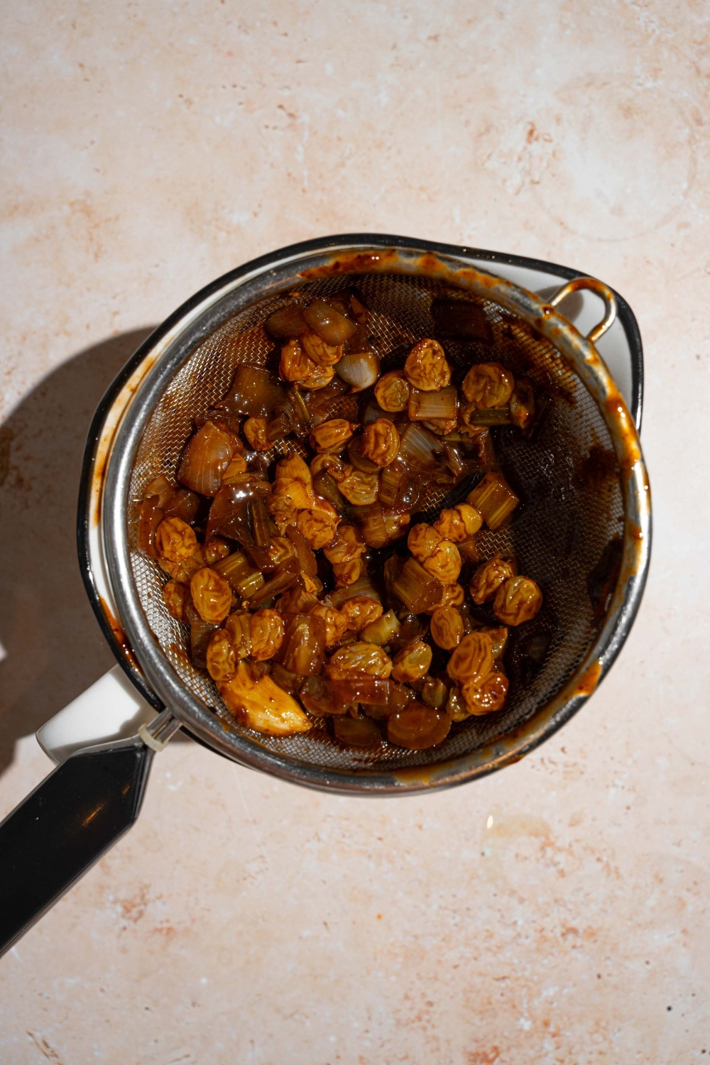 A mesh strainer with strained raisins over a stock pot. The pot is on a tan counter.