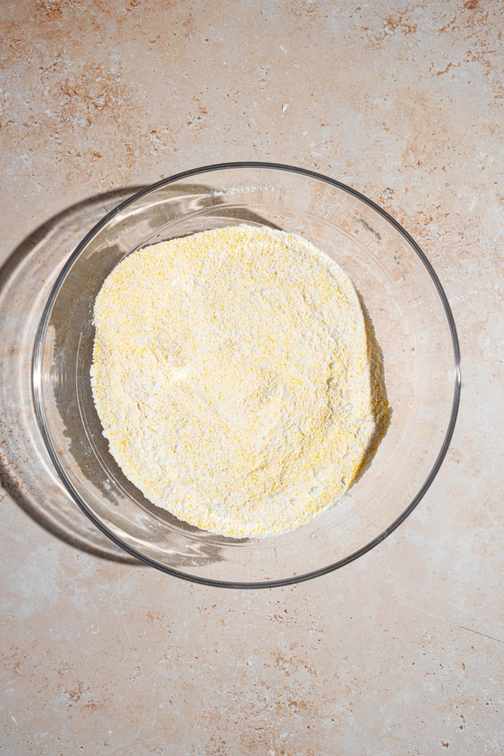 A glass bowl with dry ingredients to make cornbread. The bowl is on a tan counter.