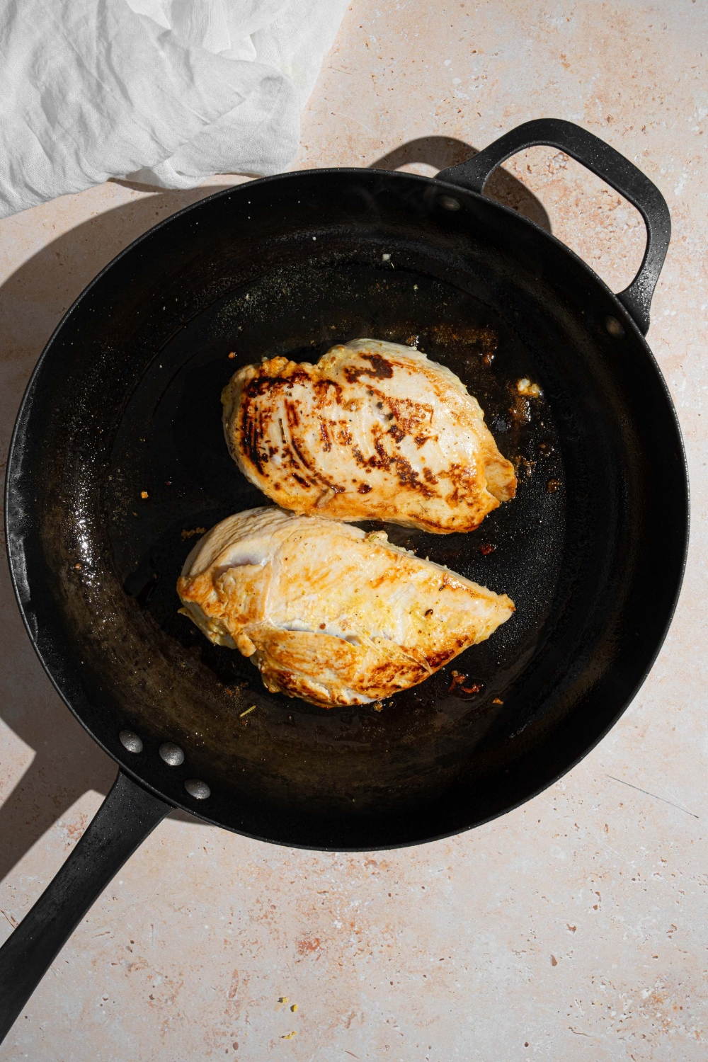 A cast iron skillet with two marinated chicken breasts cooking in oil. The skillet is on a tan counter with a white cloth napkin.