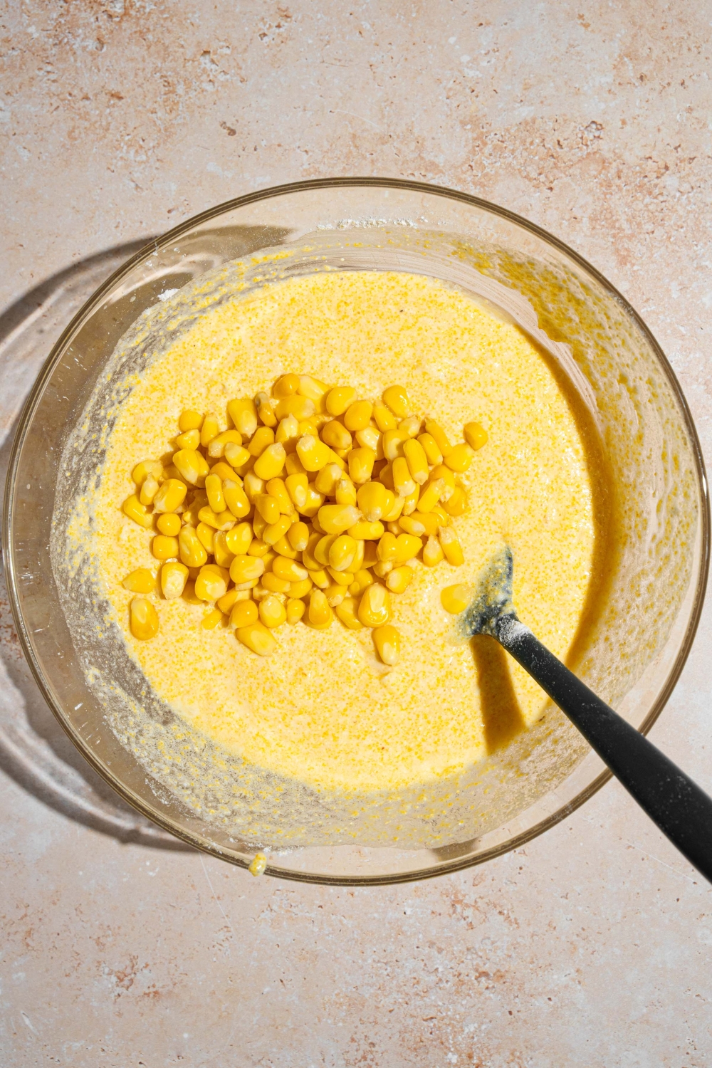 A glass bowl with a spatula stirring a cornbread batter mixture topped with corn kernels. The bowl is on a tan counter.