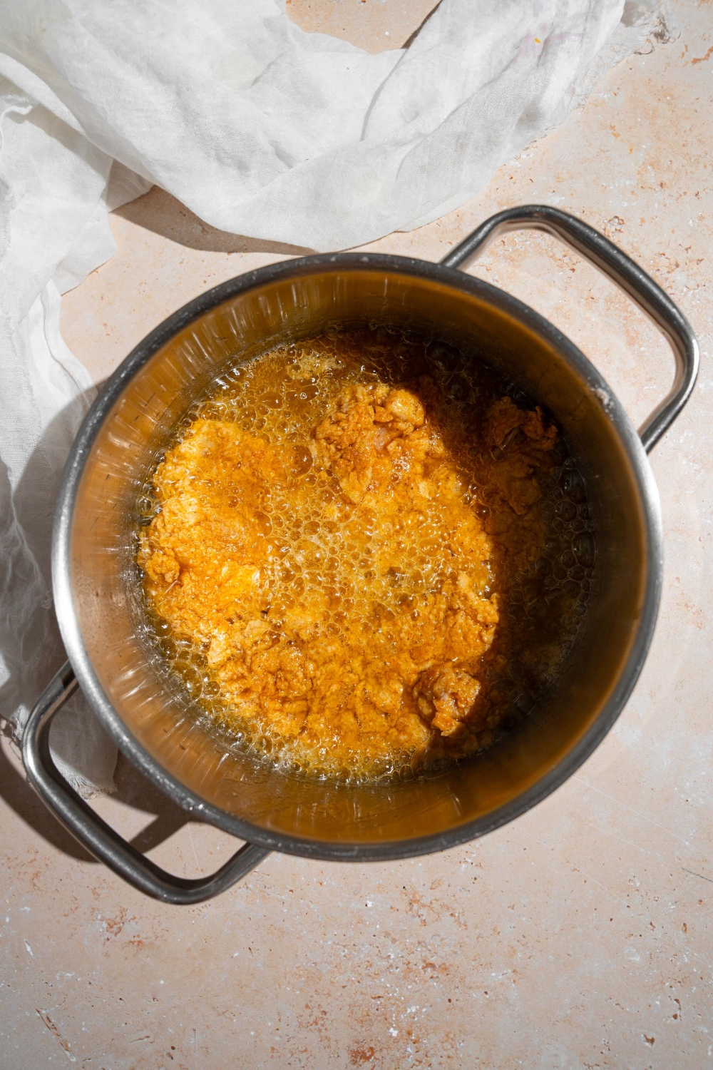 A pot with breaded chicken frying in oil. The pot is on a tan counter with a white cloth napkin.