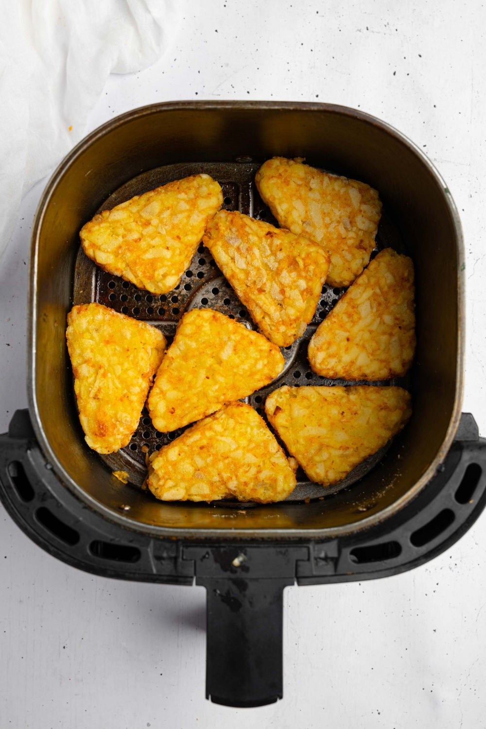 An air fryer with several hashbrowns. The air fryer is on a white counter with a white cloth napkin.