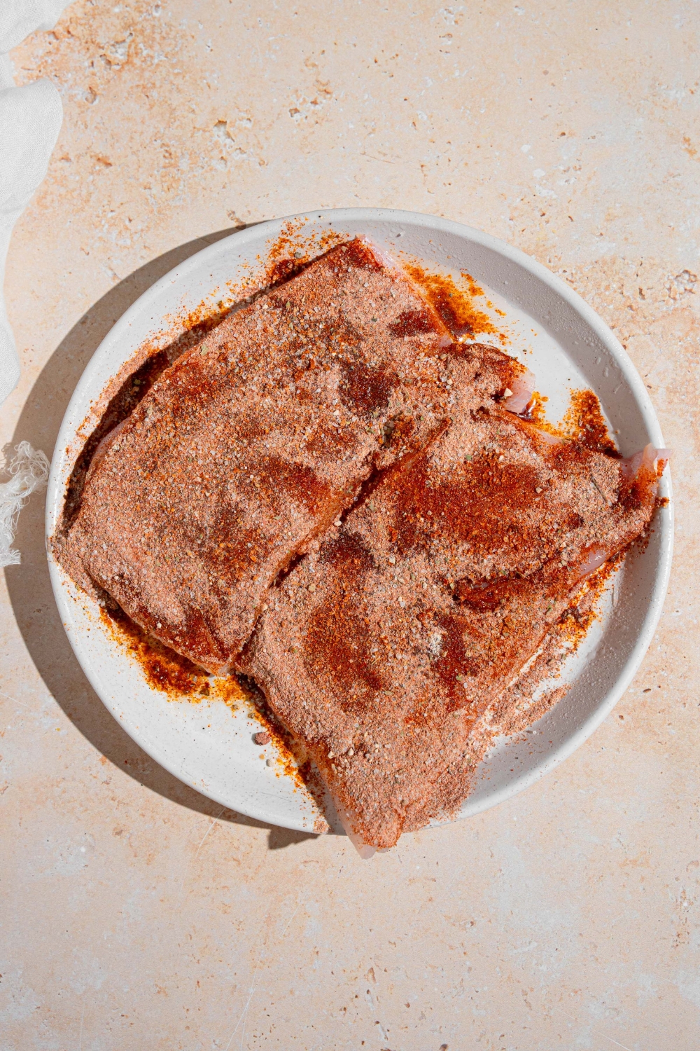 A white plate with two uncooked grouper filets coated in seasoning. The plate is on a tan counter.