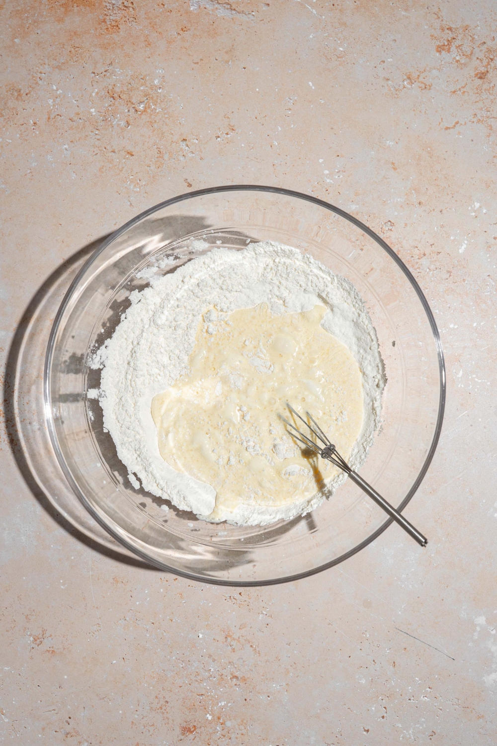 A glass bowl with a whisk combining dry and wet ingredients for pancake batter. The bowl is on a tan counter.