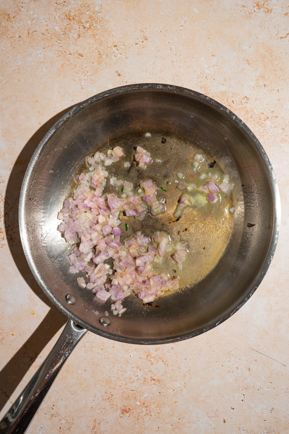 A skillet with chopped shallots sautéed in oil. The skillet is on a tan counter.