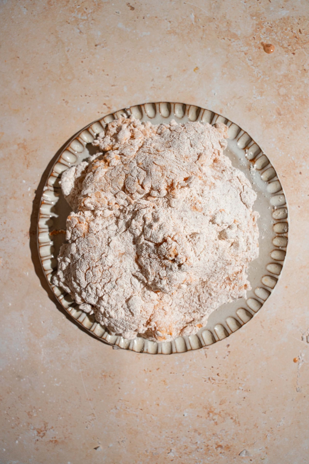 A ceramic plate with chicken breasts dredged in a seasoned coating. The plate is on a tan counter.