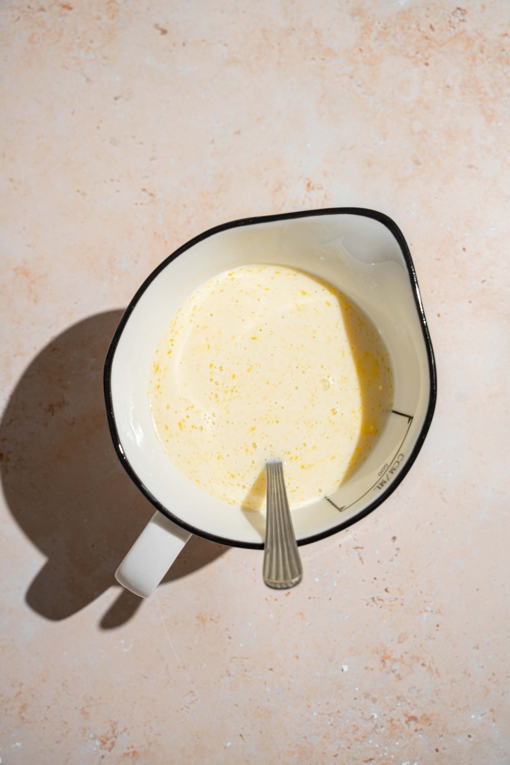A ceramic pitcher with a spoon stirring pancake batter mix. The pitcher is on a tan counter.