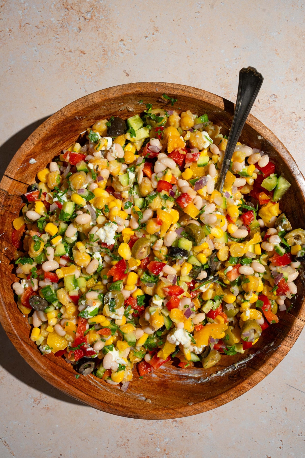 A wooden bowl with a spoon mixing ingredients to make Mediterranean salad including navy beans, chickpeas, bell peppers, cucumber, corn, and olives with a lemon vinaigrette. The bowl is on a tan counter.