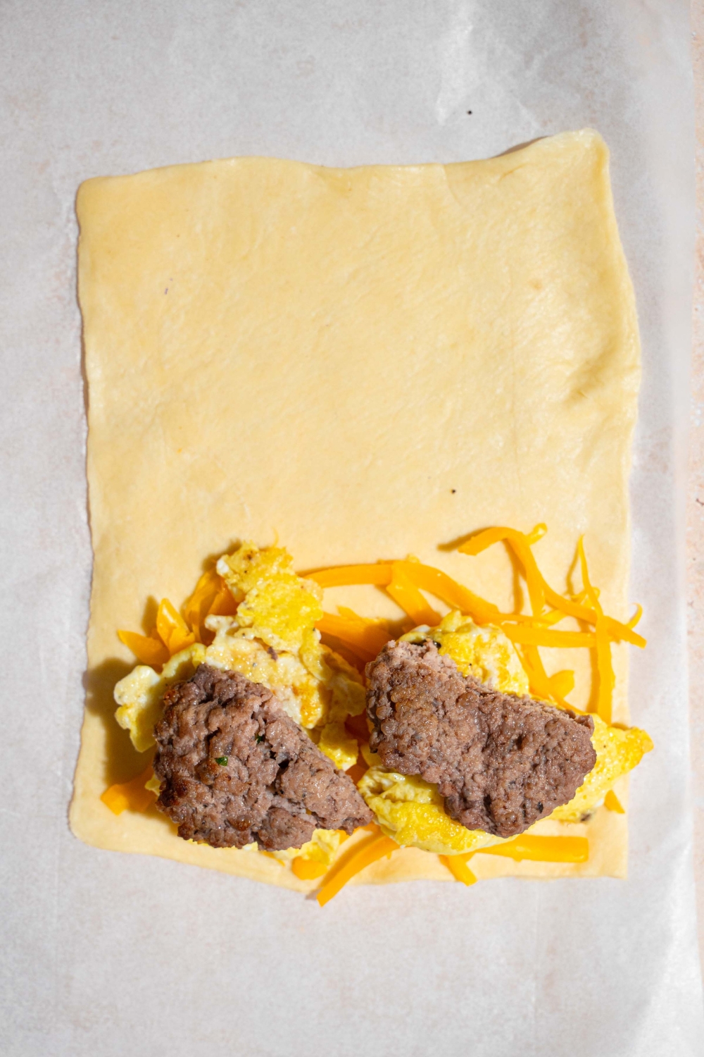 A sheet of puff pastry with shredded cheese, scrambled eggs, and breakfast sausage patties on the bottom half of the sheet. The pastry is on a sheet of parchment paper.