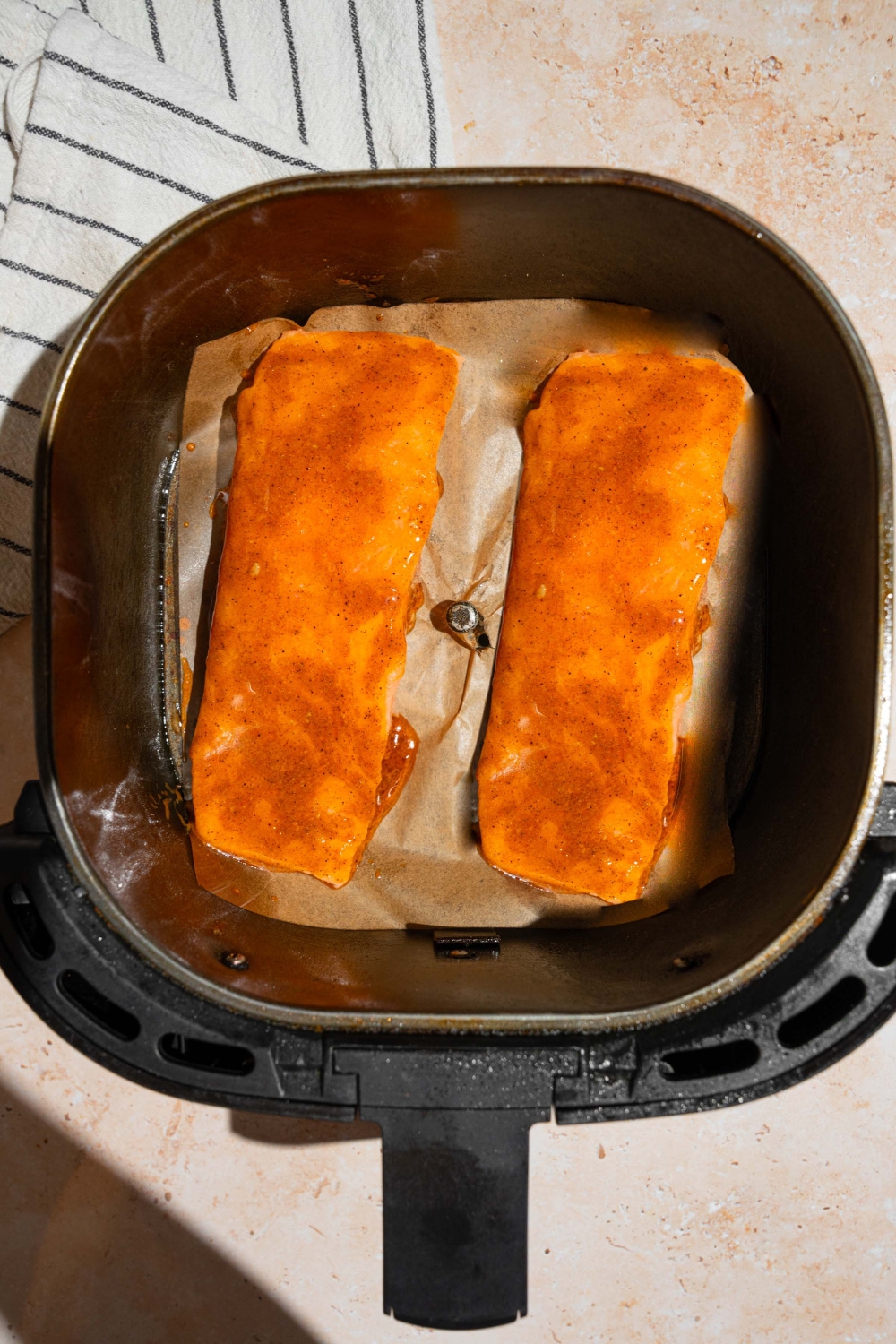 An air fryer with marinated frozen salmon filets. The air fryer is on a tan counter with a white striped napkin.