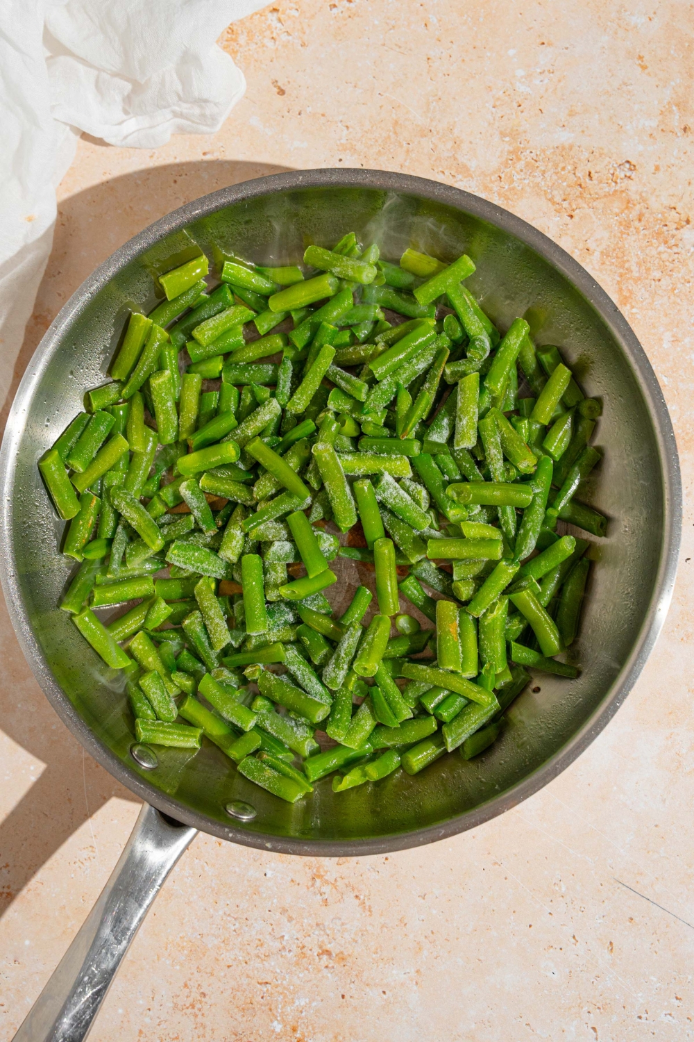 A skillet with frozen green beans. The skillet is on a tan counter with a white cloth napkin.