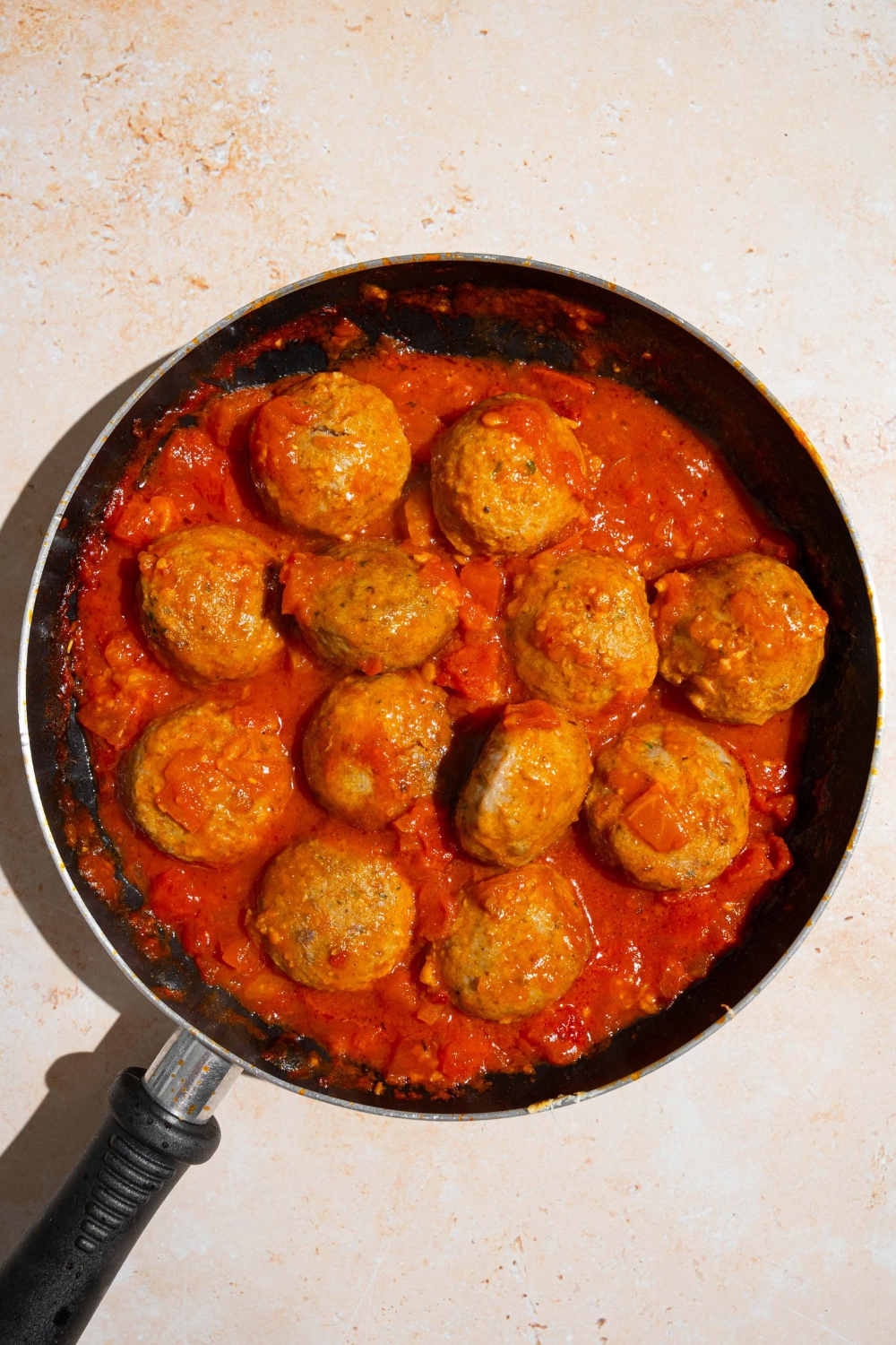 A skillet with several frozen meatballs cooking in tomato sauce. The skillet is on a tan counter.