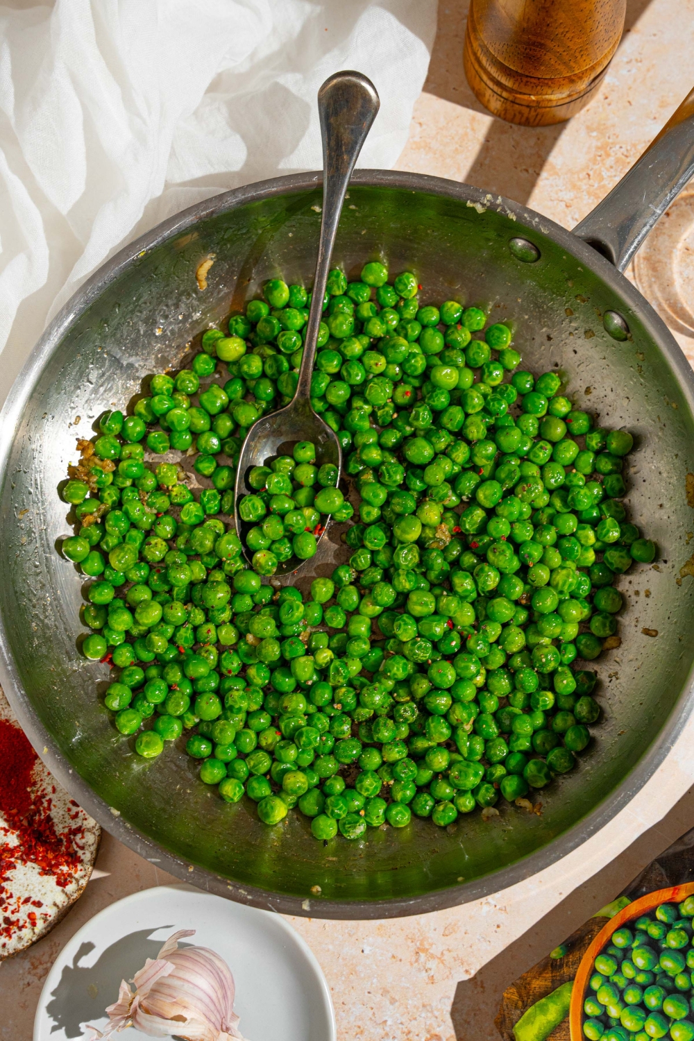 A skillet with cooked frozen peas tossed in seasonings. There is a spoon scooping the peas. The skillet is on a tan counter with a white cloth napkin and small bowl of seasonings.