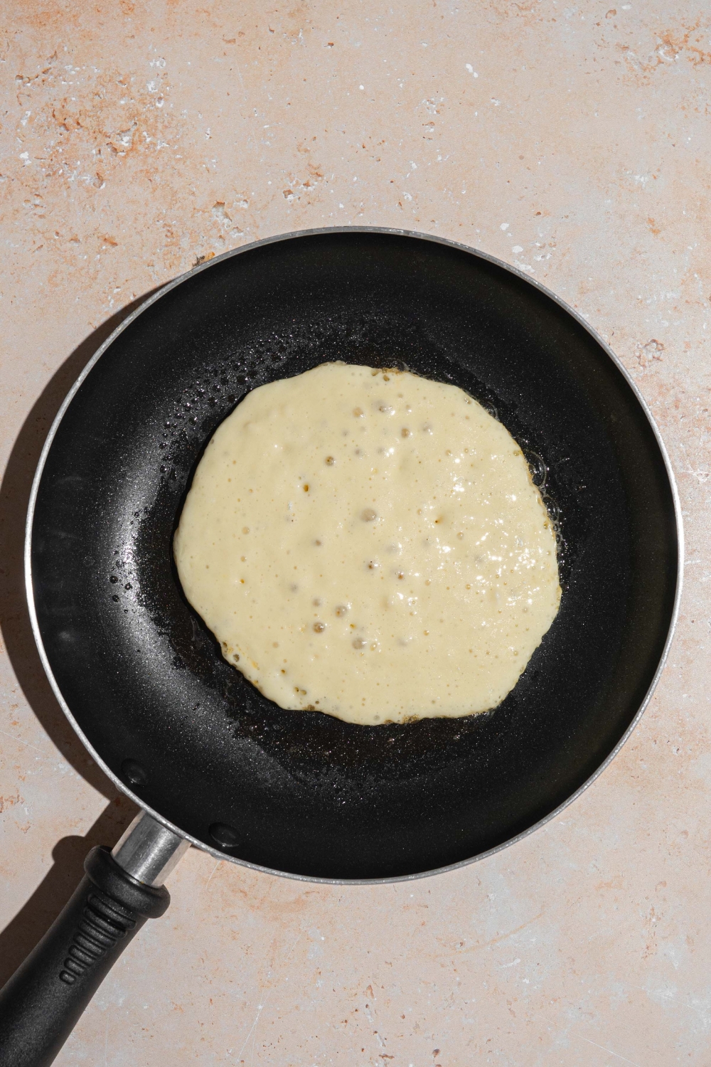A skillet with a pancake cooking in butter. The skillet is on a tan counter.