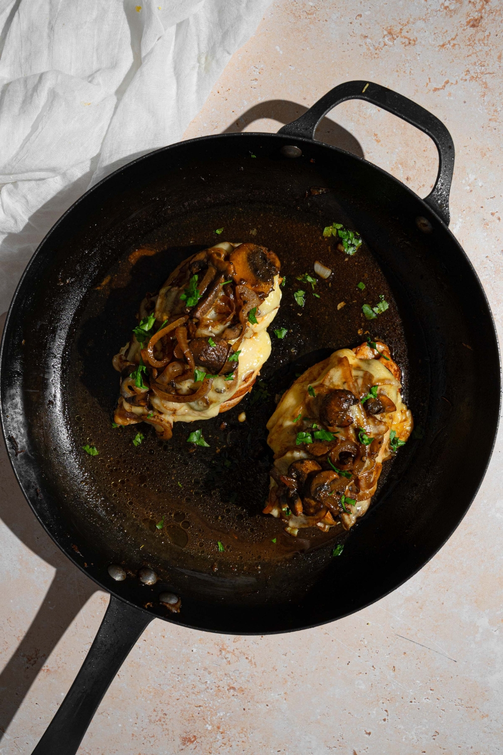 Two Texas Roadhouse smothered chicken breasts on a skillet in juices and garnished with fresh parsley. The skillet is on a tan counter with a white cloth napkin.