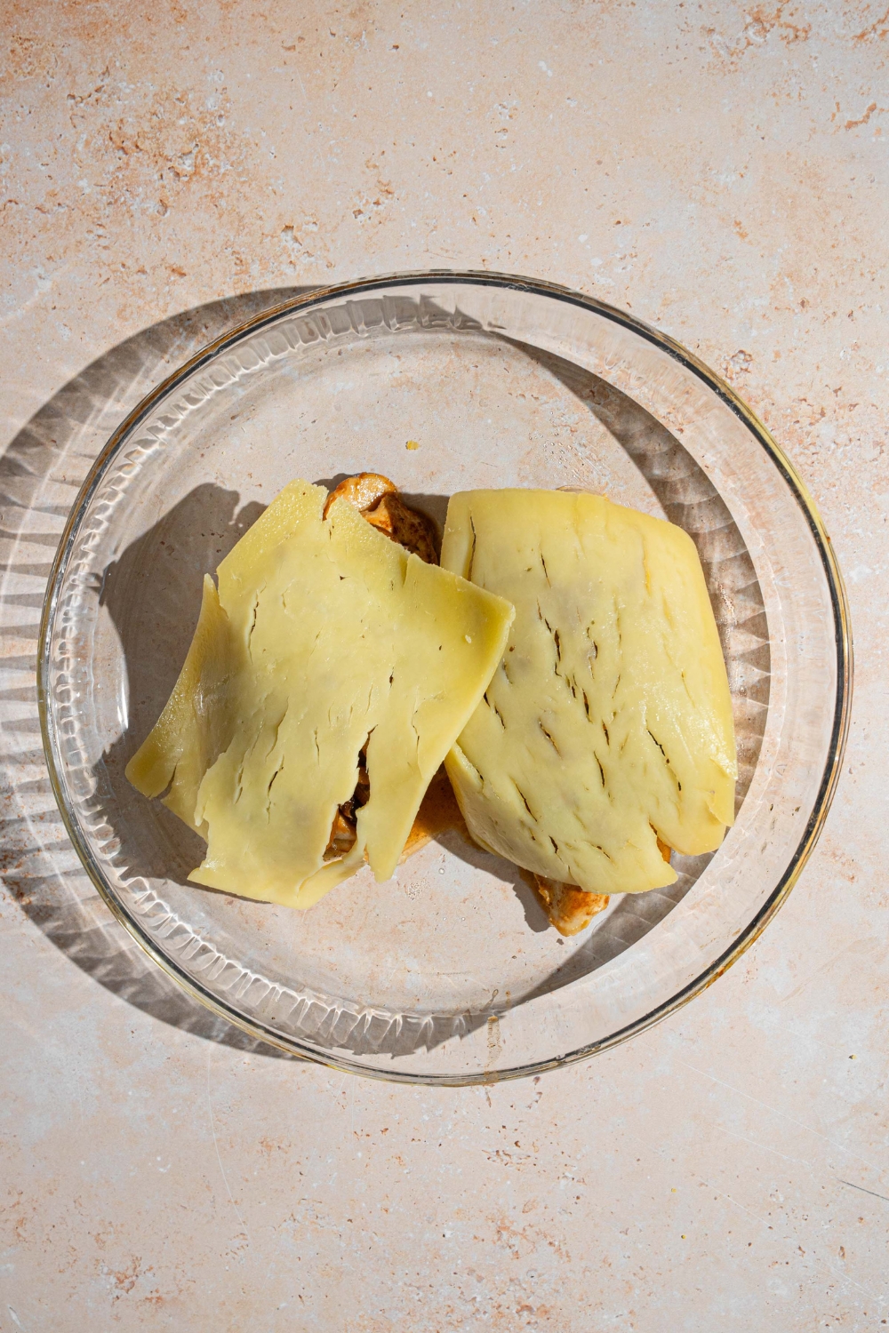 Two cooked chicken breasts topped with slices of monterey jack cheese. The chicken is on a glass baking dish. The dish is on a tan counter.