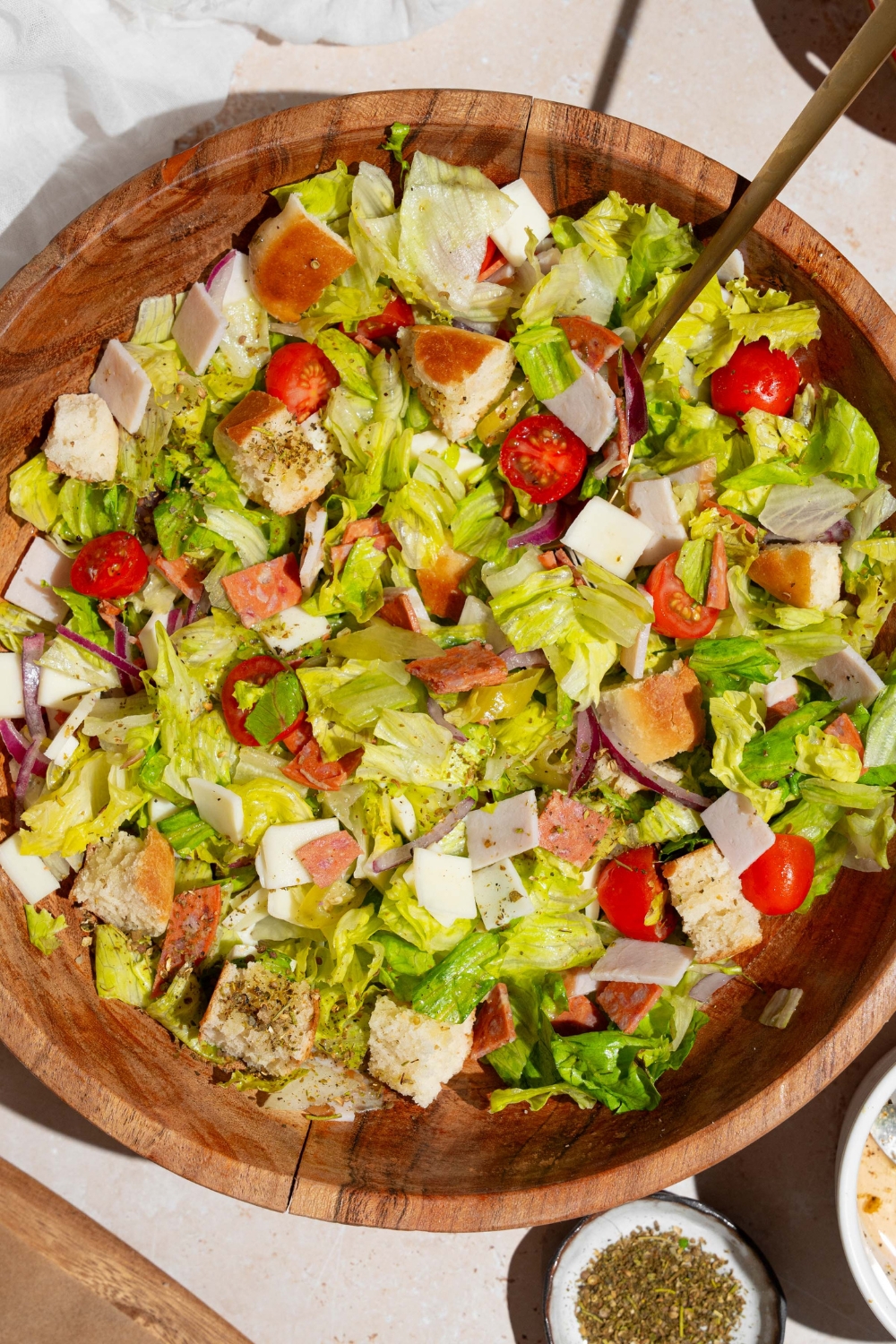 A wooden salad bowl of copycat Jersey Mike's sub in a tub including mixed lettuce, tomatoes, provolone cheese, diced meats, and bread croutons. There is a fork in the salad. The bowl is on a tan counter with a small ramekin of seasonings and dressing.