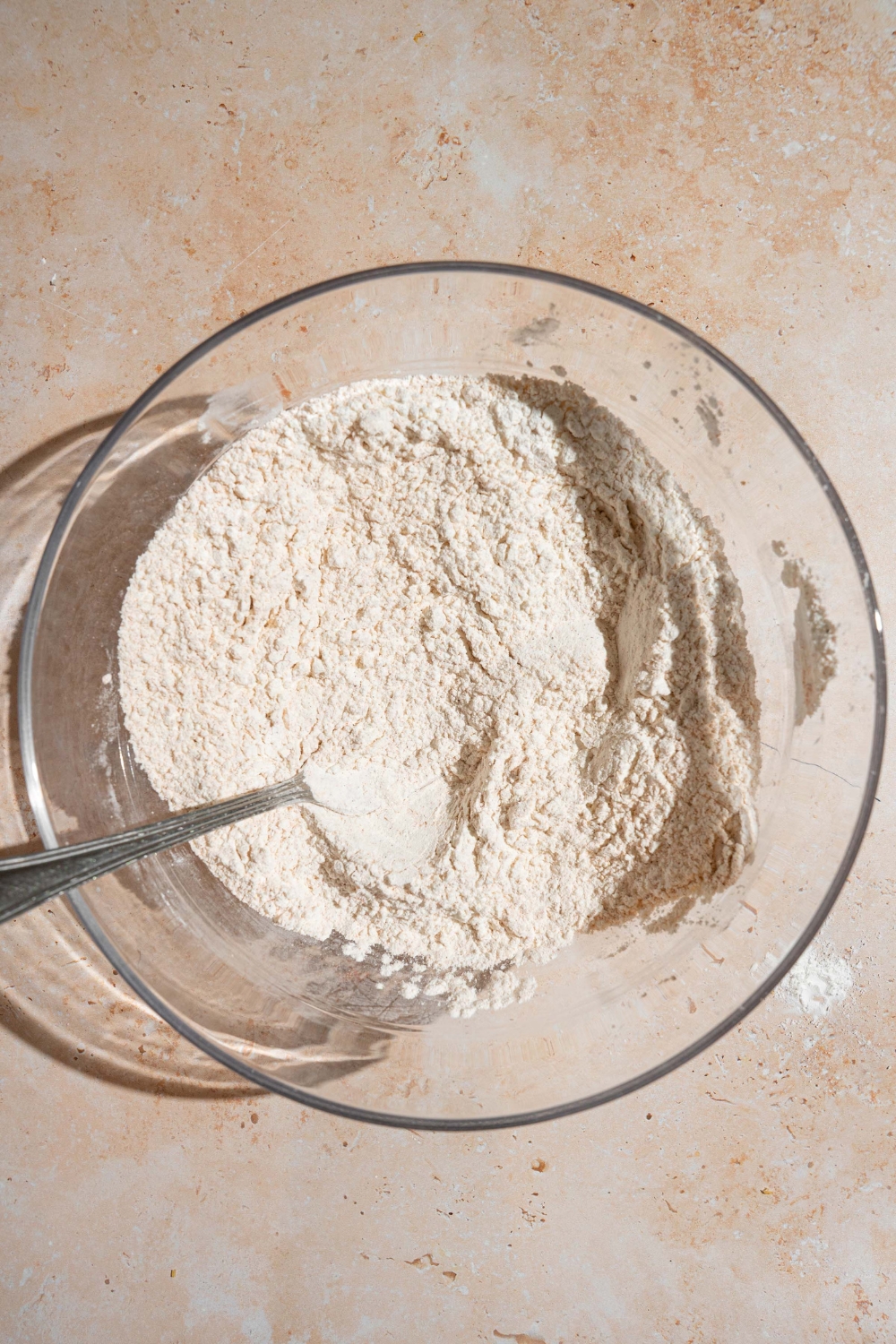 A glass bowl with a spoon mixing a seasoned flour mixture. The bowl is on a tan counter.