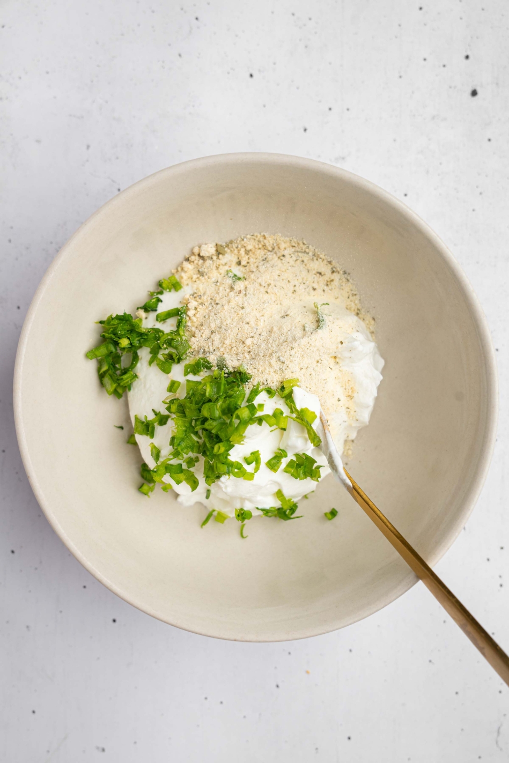 A ceramic bowl with a spoon mixing sour cream, ranch seasoning, and chives. The bowl is on a white counter.