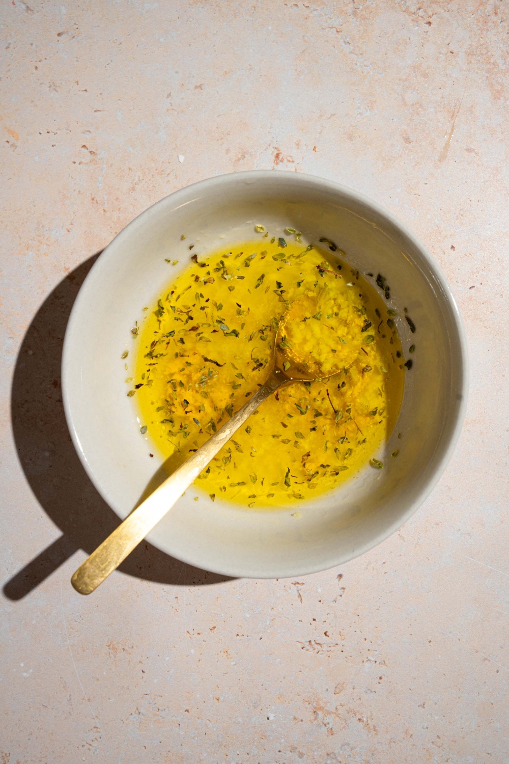 A small white bowl with a spoon stirring a lemon vinaigrette dressing. The bowl is on a tan counter.