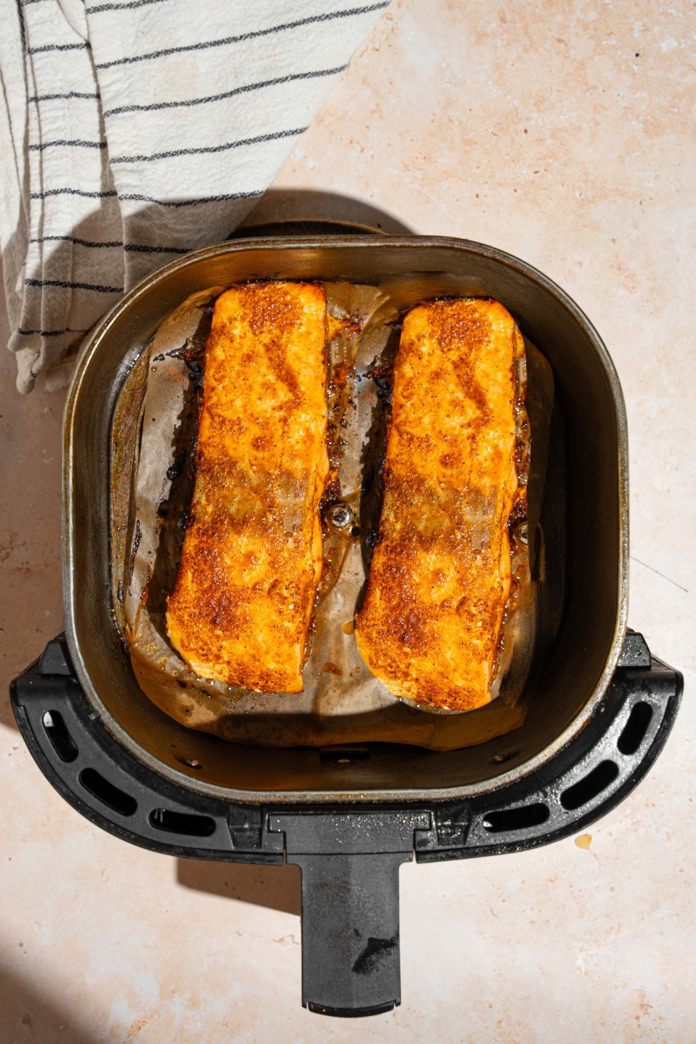 An air fryer with two marinated salmon fillets. The air fryer is on a tan counter with a white striped napkin.