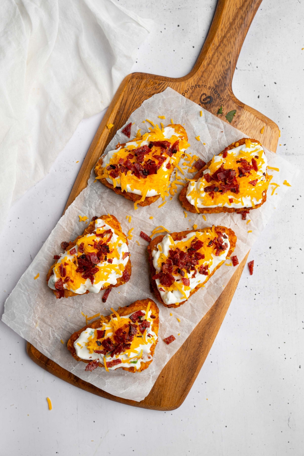 A wooden board lined with parchment paper with crispy hashbrowns layered with sour cream, cheddar cheese, and crumbled bacon. The board is on a white counter with a white cloth napkin.