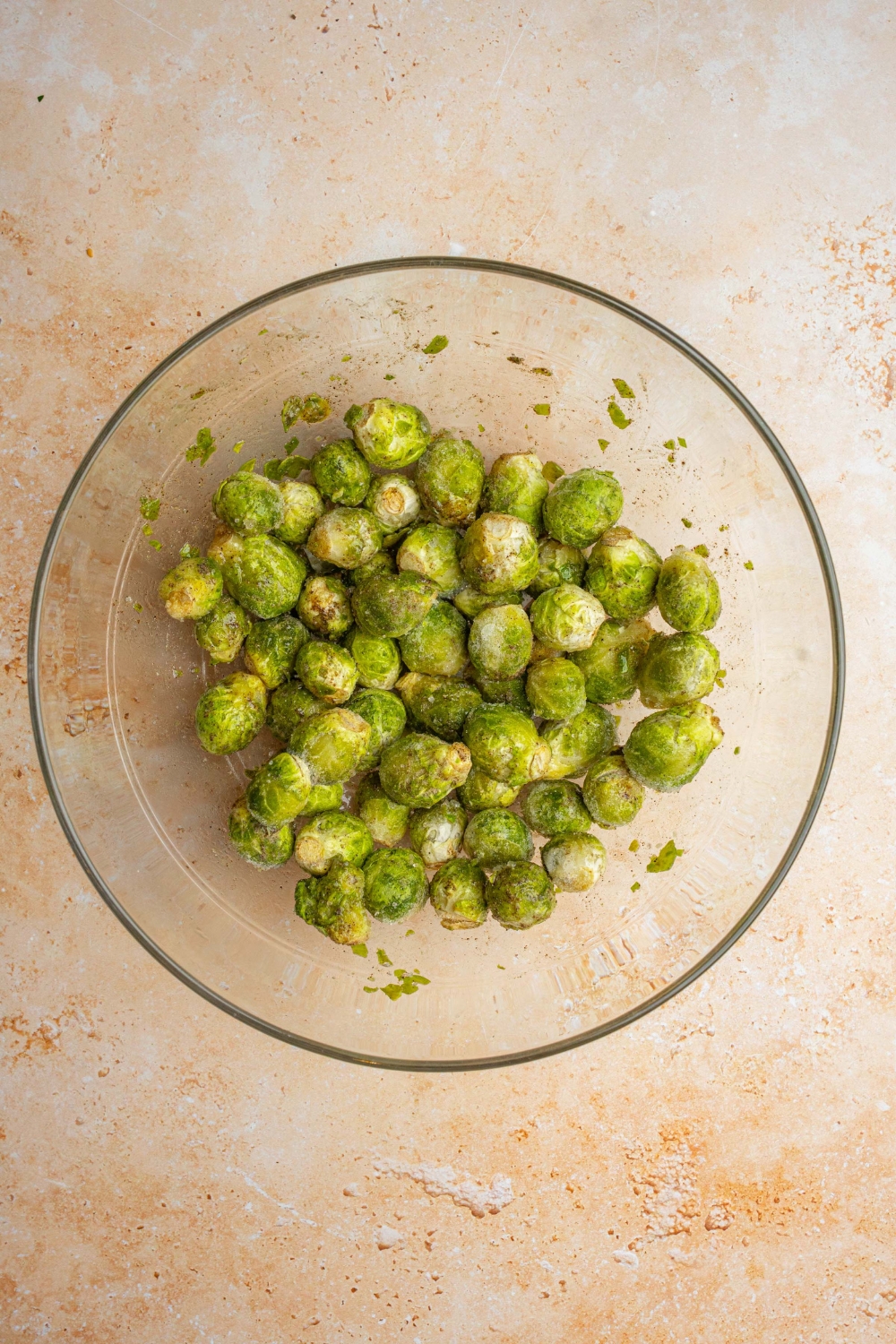 A glass bowl with frozen brussel sprouts drizzled with olive oil. The bowl is on a tan counter.