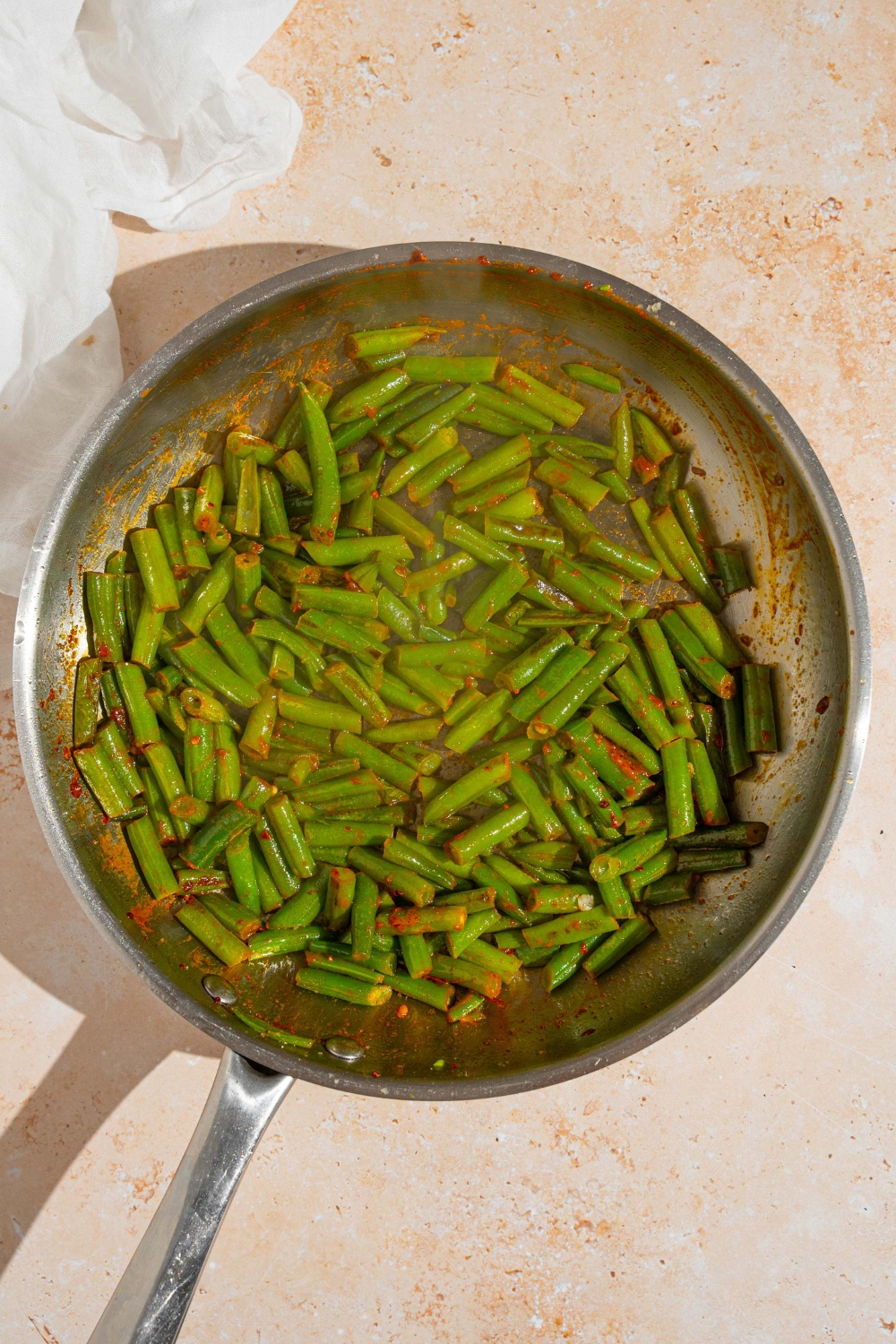 A skillet with frozen green beans sautéing in oil with seasonings. The skillet is on a tan counter with a white cloth napkin.