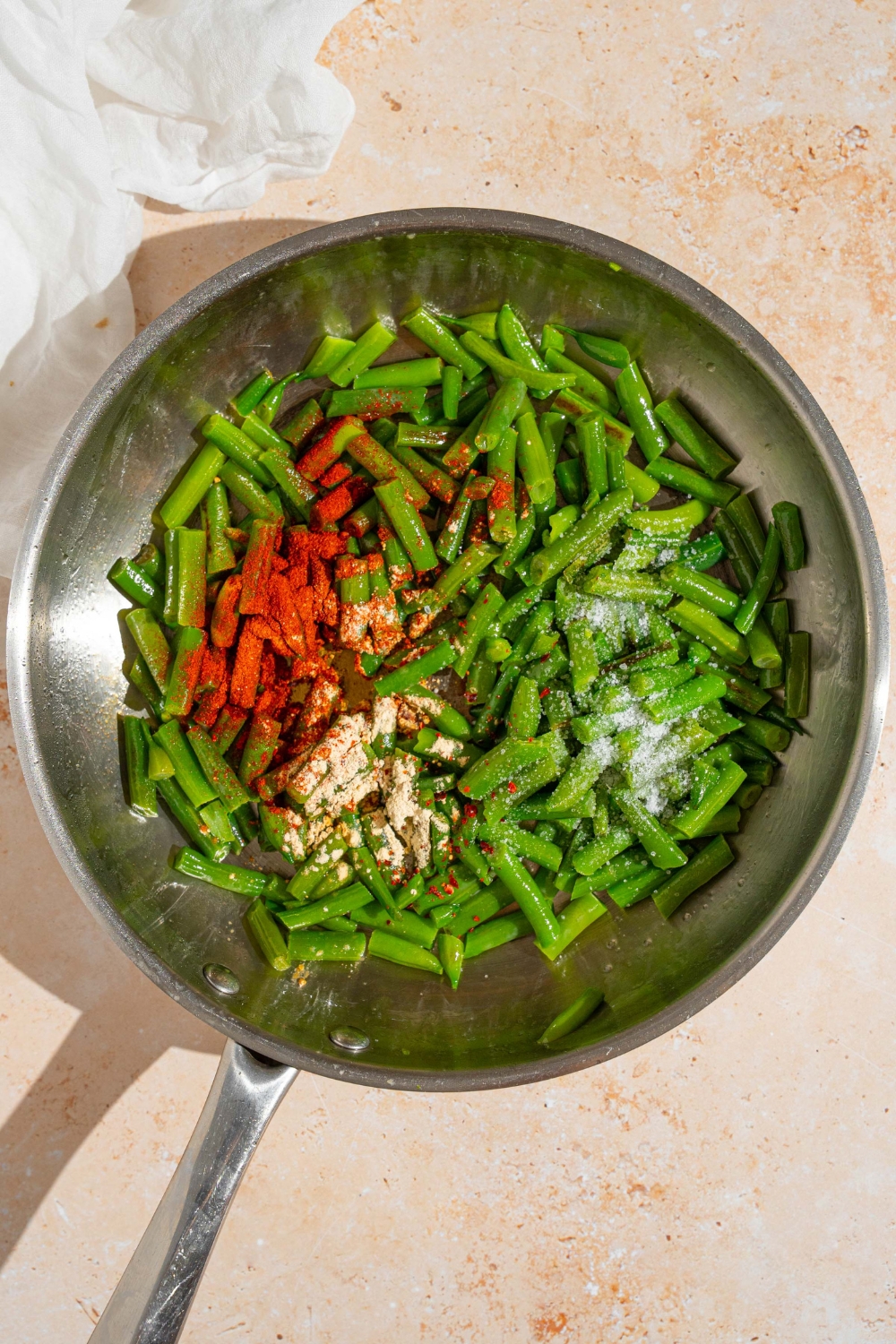 A skillet with frozen green beans topped with seasonings including paprika, garlic powder, salt, and pepper. The skillet is on a tan counter.