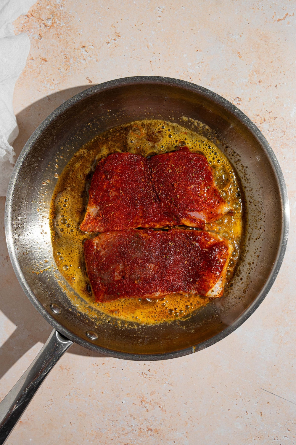 A skillet with two seasoned groupers cooking in butter. The skillet is on a tan counter.