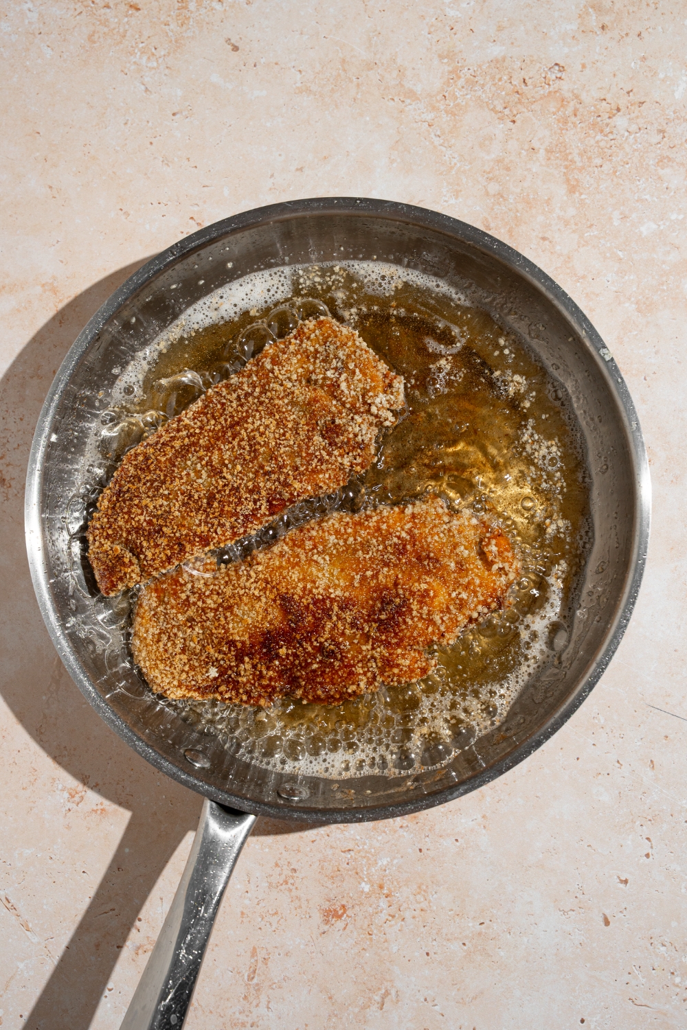 A skillet with two breaded chicken cutlets frying in oil. The skillet is on a tan counter.