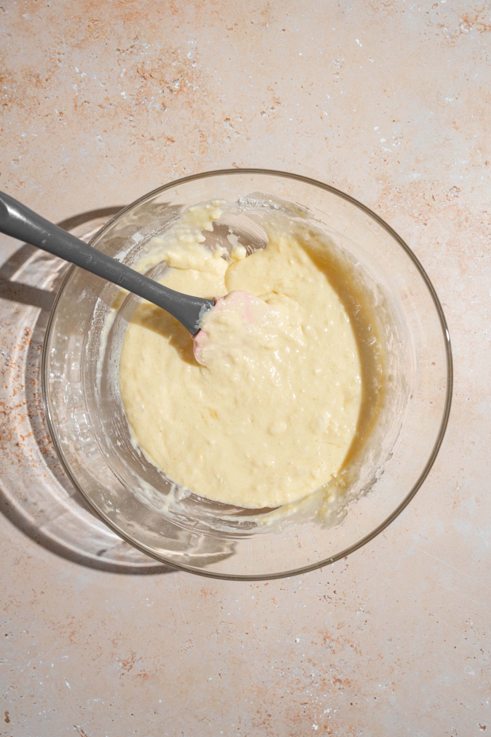 A glass bowl with a spatula mixing pancake batter. The bowl is on a tan counter.