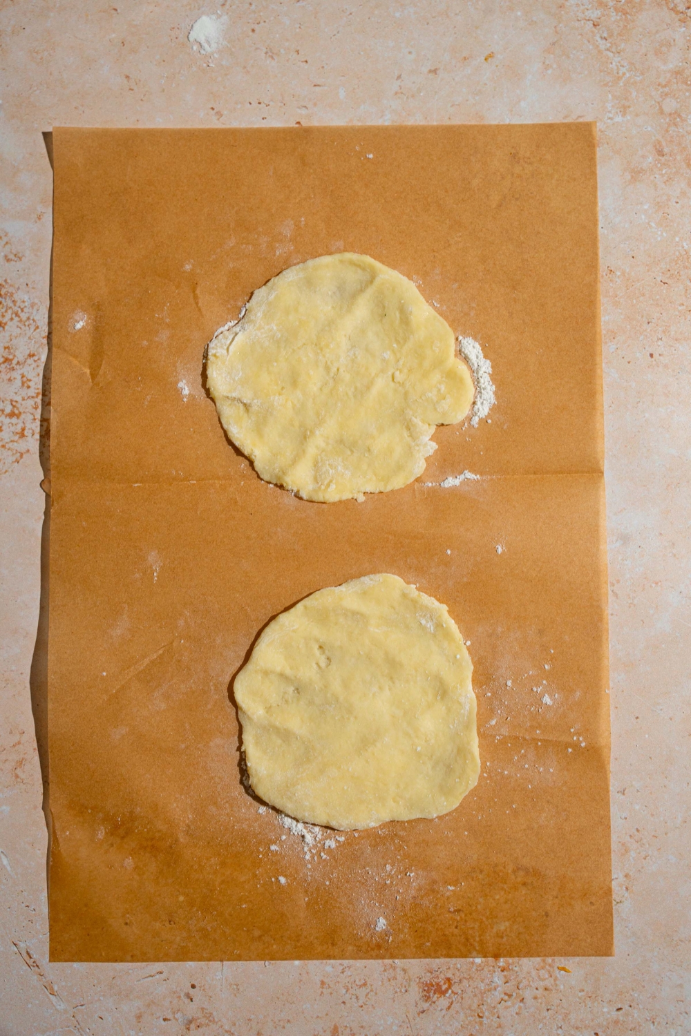 A piece of parchment paper dusted with flour with two pieces of shaped biscuit dough. The paper is on a tan counter.