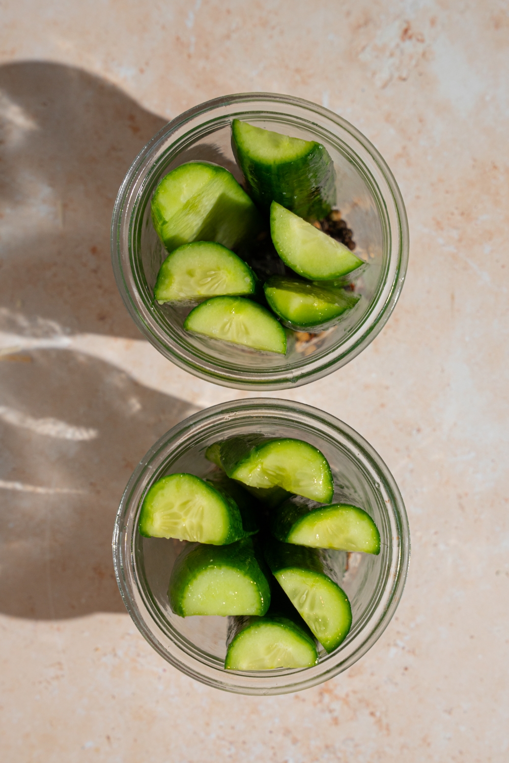 Two glass jars with medium pickling cucumber spears on top of picking seasonings. The jars are on a tan counter.